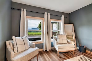 Una habitación con dos sillones, un ventanal con cortinas y vistas a zonas verdes, y suelo de madera.