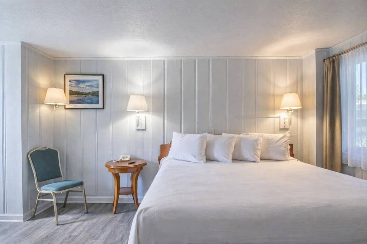 A hotel room with a bed, two bedside lamps, a small table with a chair, and a painting on the wall.
