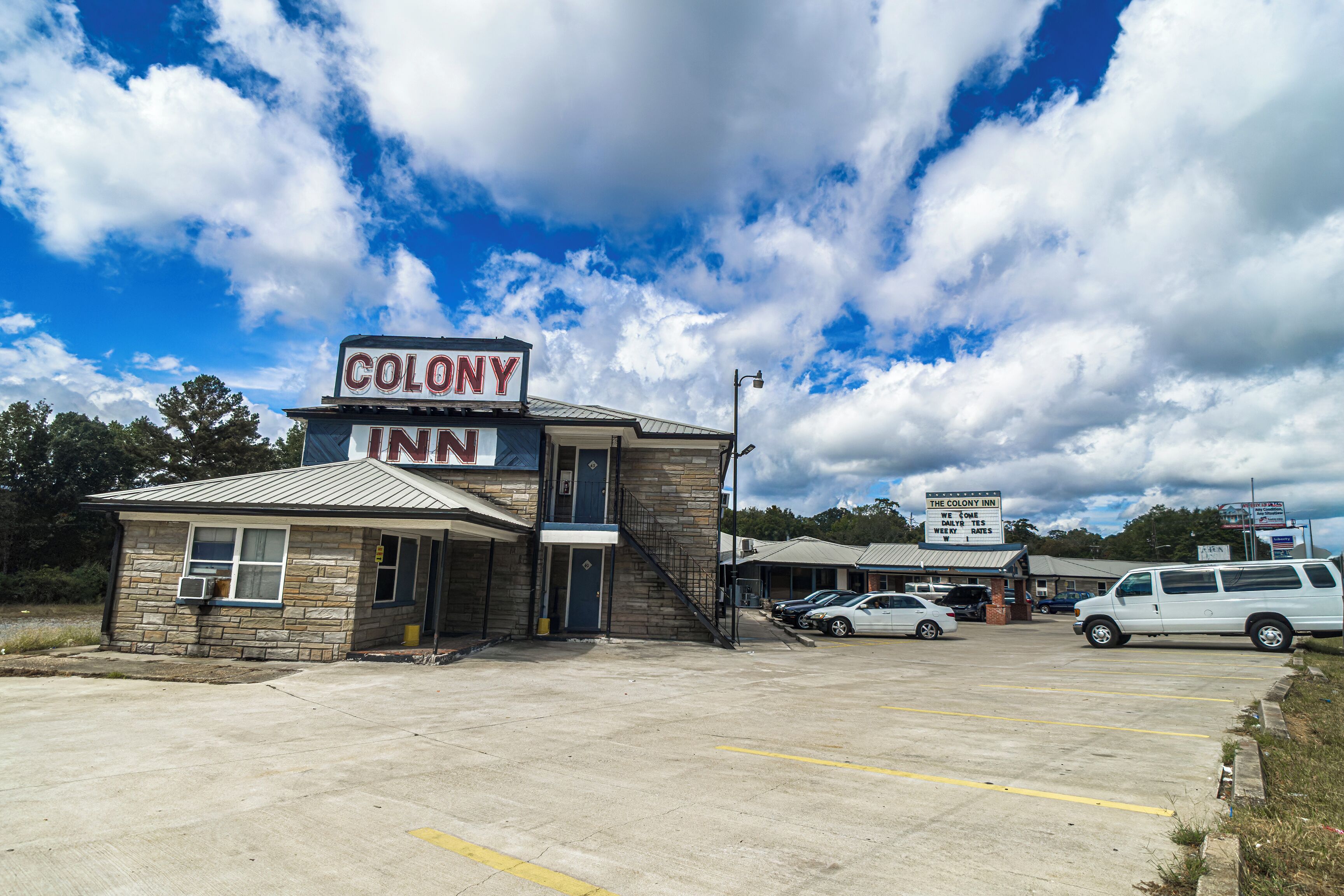 Un bâtiment à deux étages, sur lequel on peut lire l’inscription « Colony Inn ». Des voitures sont garées devant, et le ciel est dégagé, avec quelques nuages au-dessus.