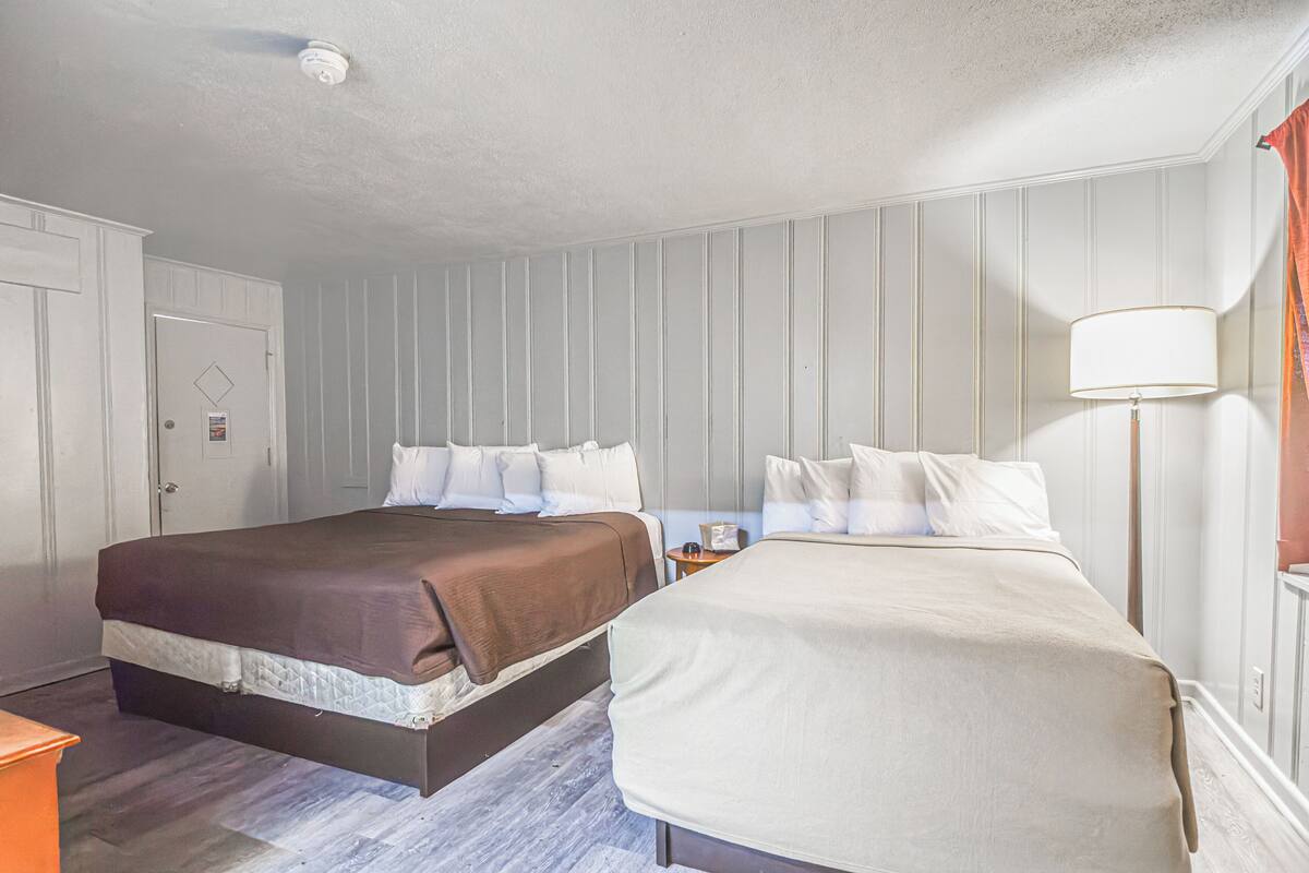 A hotel room with two beds, a nightstand, a lamp, and a door.