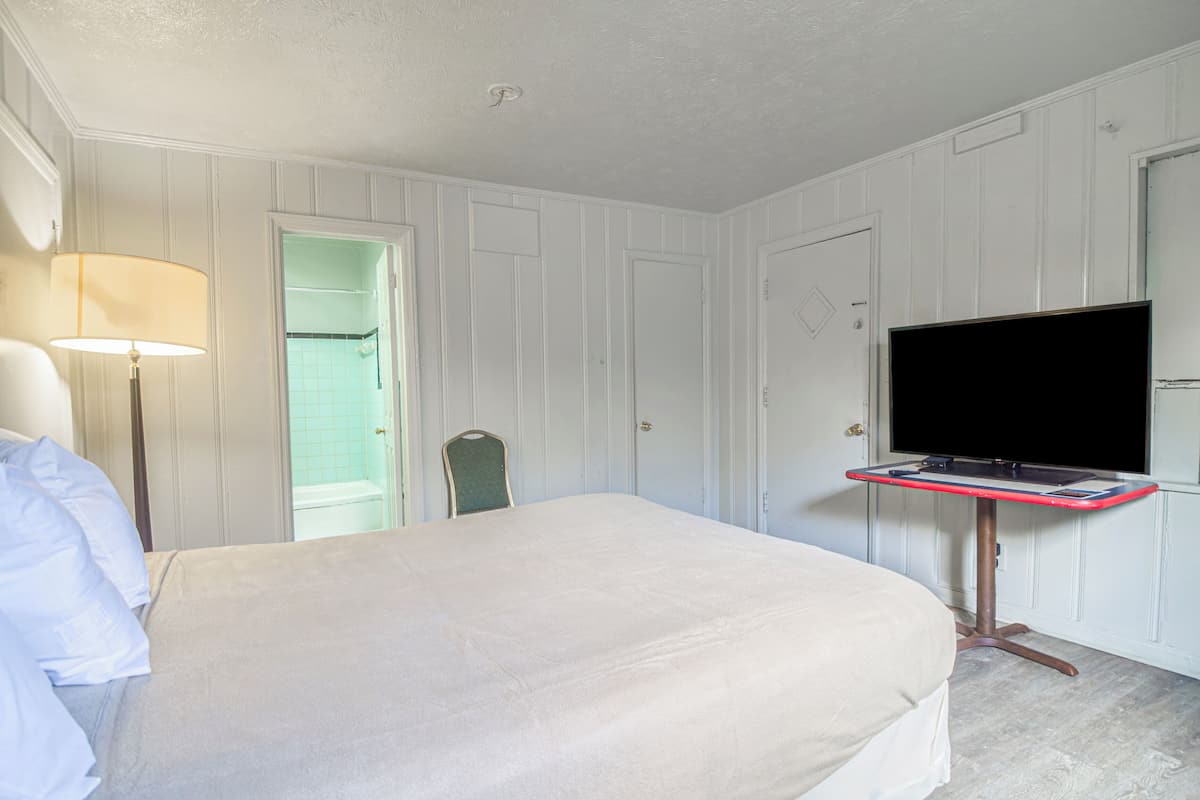 A hotel room with a bed, a desk with a flat-screen TV, a lamp, and a door leading to a bathroom.