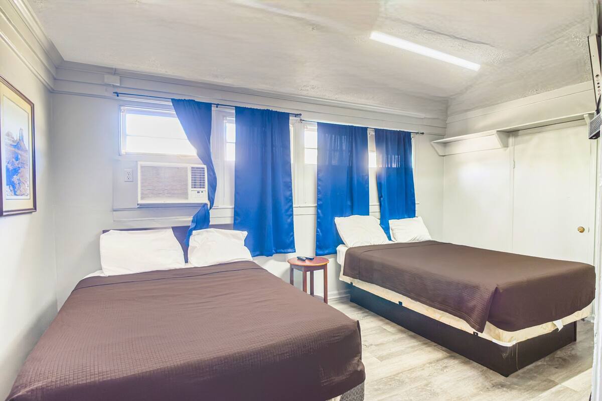 A room with two beds, a small table, a window with curtains, and an air conditioning unit.