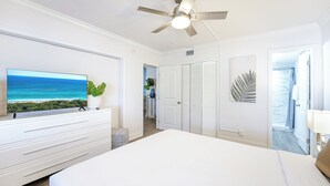 A bedroom with a large bed, a ceiling fan, a TV displaying a beach scene, and a view of the ocean through a window. - Boca Waterfront • Walk to Beach • Pool & Hot Tub • Gym • Parking (Boca Raton)