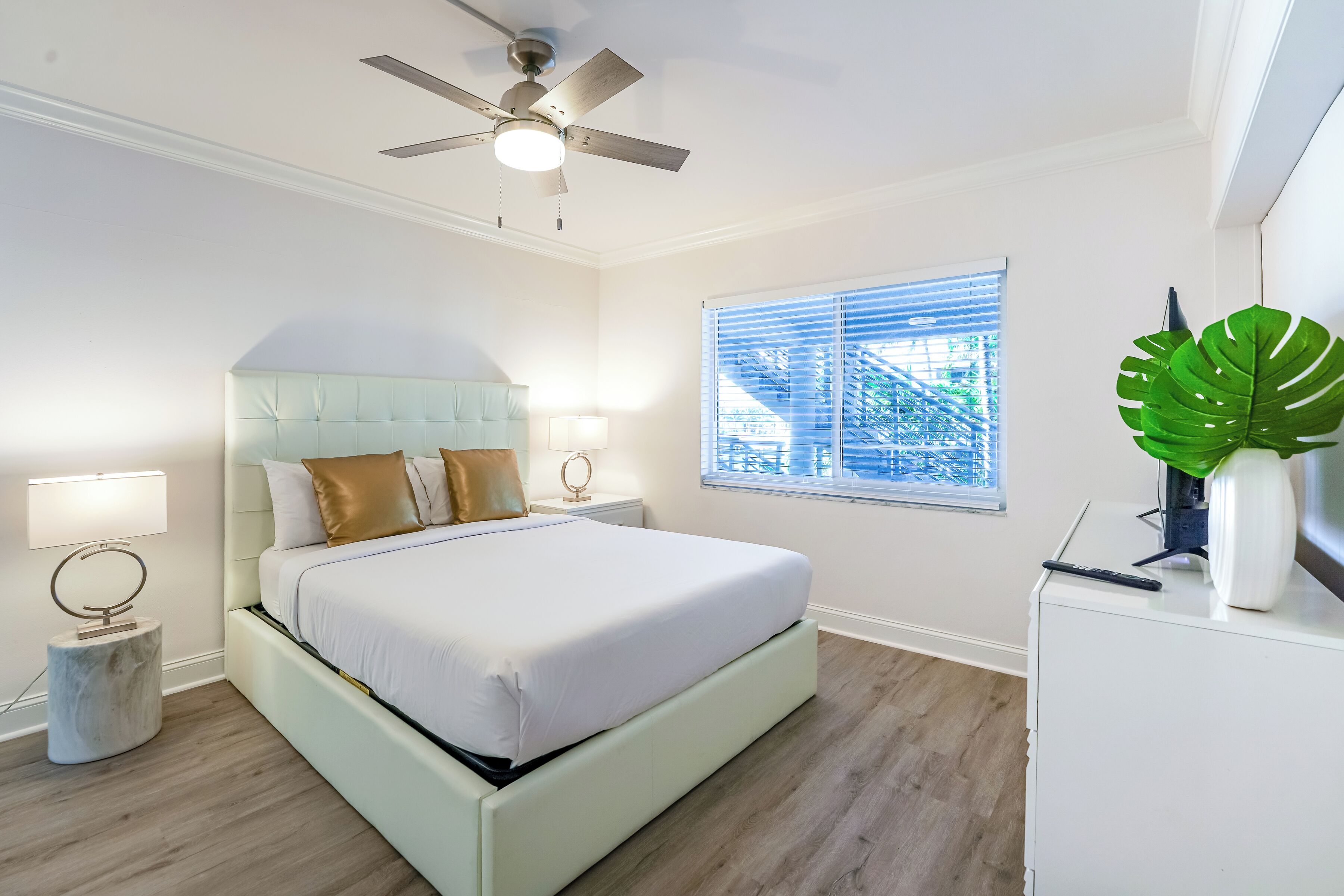 Quarto com cama, ventilador de teto, abajures, janela com persianas e uma planta em uma cômoda.