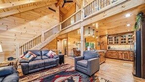 Interior espacioso de una cabaña de madera, con sala de estar, cocina y una escalera que sube a un nivel superior.