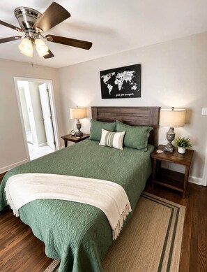 A bedroom with a bed, two bedside tables, a world map artwork, a ceiling fan, and a door.