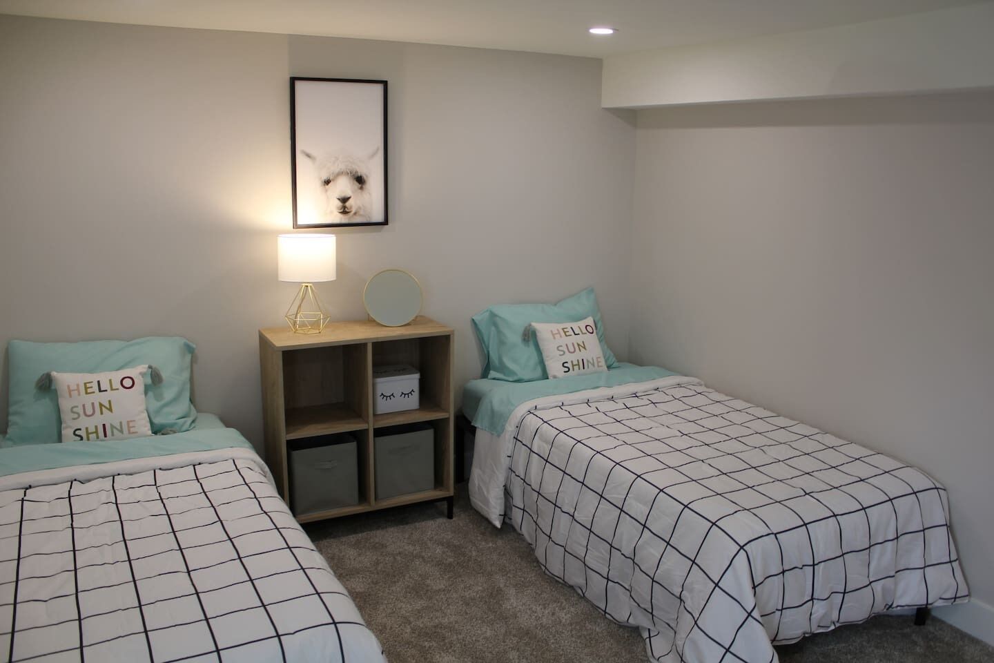 Un dormitorio con dos camas, cada una con una colcha a cuadros y almohadas con el texto 'Hello Sun Shine'. Hay una mesita de noche con una lámpara, un espejo y cajas de almacenamiento.