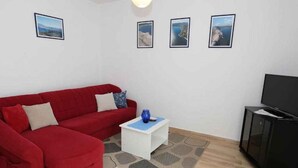 Ein Wohnzimmer mit einem roten Sofa, einem kleinen weißen Beistelltisch und einer schwarzen Vitrine mit einem Fernseher. An der Wand hängen drei gerahmte Bilder.