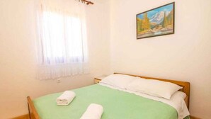 Ein Einzelbett mit einer grünen Bettdecke und weißen Kissen, ein Gemälde einer Berglandschaft an der Wand und ein Fenster mit durchscheinenden Vorhängen.