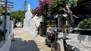 Uma rua estreita com edifícios brancos, uma motocicleta estacionada na lateral e uma árvore com flores roxas.
