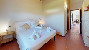 A bedroom with a wooden ceiling, a bed with white linens, a bedside table with a lamp, and a view into another room with a dining area.