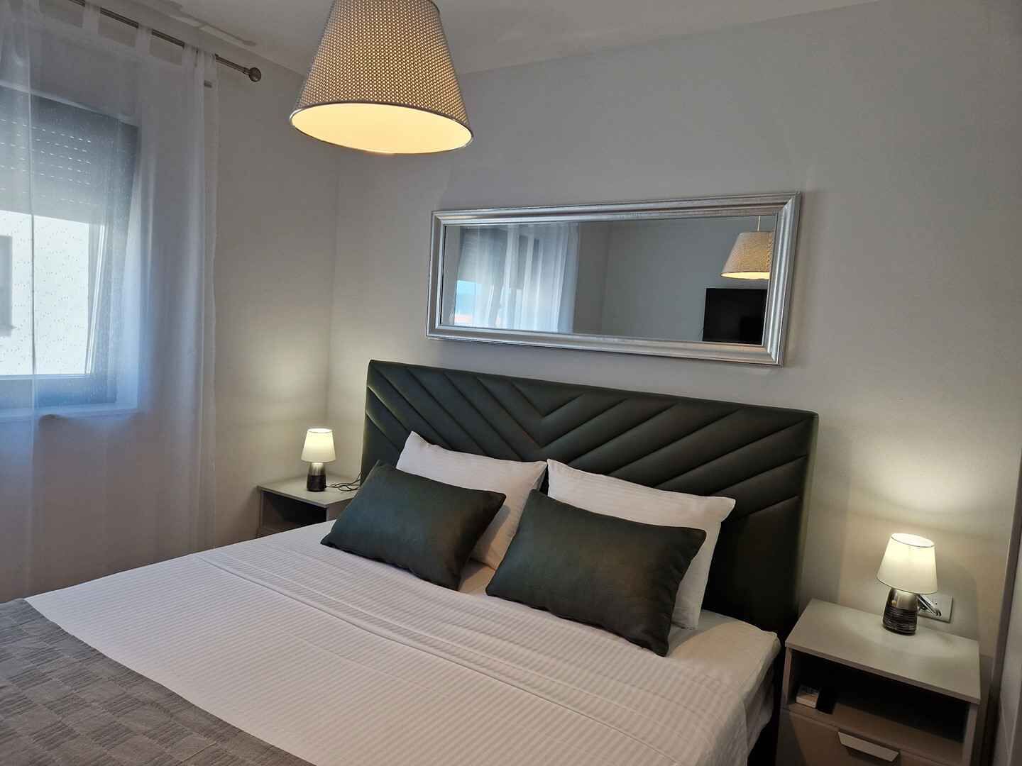 Ein ordentlich bezogenes Bett mit Kopfteil, zwei Nachttische mit Lampen, ein großer Spiegel an der Wand und ein Fenster mit Vorhängen.