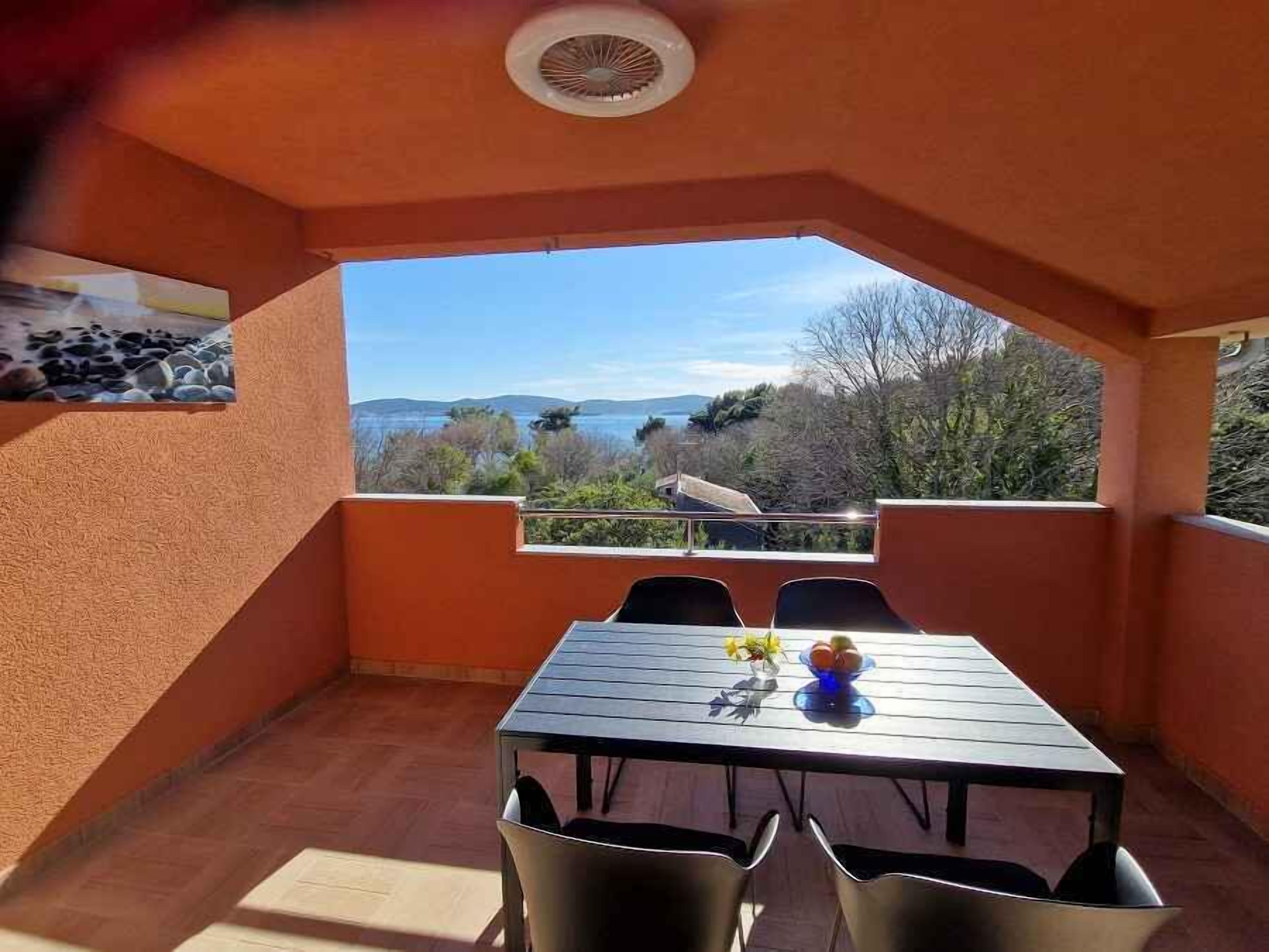 A small outdoor balcony with a table and chairs, a view of the sea and hills, and a ceiling fan.