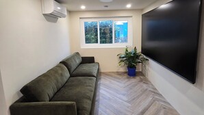 Un salon moderne avec un canapé d’angle, un téléviseur à écran plat fixé au mur, une fenêtre donnant sur un espace verdoyant et un climatiseur fixé au plafond.