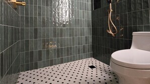Une salle de bain moderne avec des murs recouverts de carreaux foncés, des toilettes blanches et une douche dotée d’une robinetterie dorée.