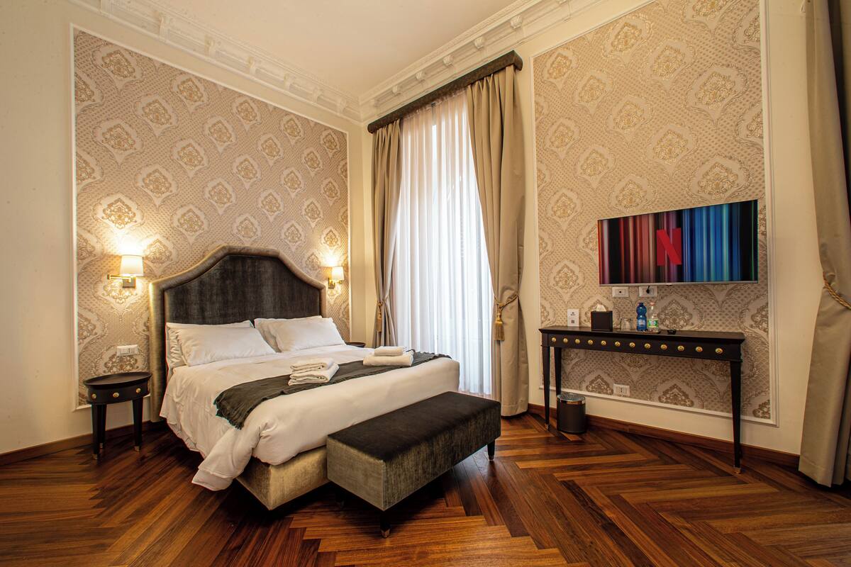 A well-appointed bedroom with a large bed, bedside lamps, a nightstand, a bench, a small table, and a wall-mounted TV.
