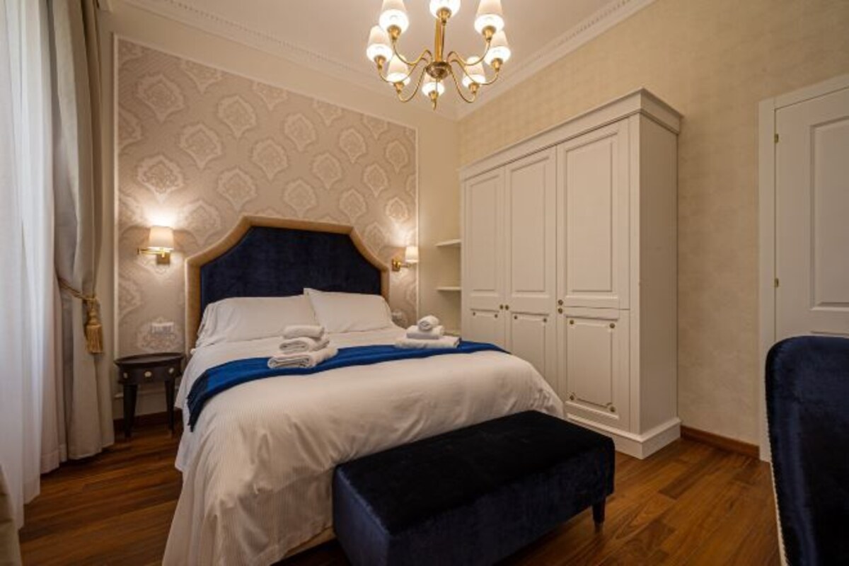 A hotel room with a large bed, a wardrobe, a bench, and a chandelier.