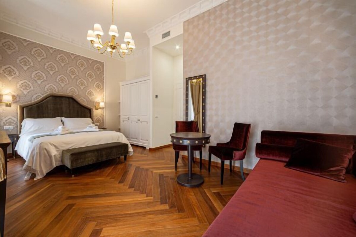 A hotel room with a wooden herringbone floor, a large bed with a dark headboard, a round table with chairs, and a red sofa.