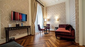 Une chambre d’hôtel avec un parquet en bois disposé en chevrons, un canapé en velours rouge, un téléviseur fixé au mur, un bureau avec une lampe et un papier peint à motifs.