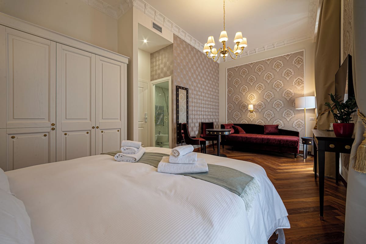 A hotel room with a large bed, a sitting area with a red sofa, a small table, and a chandelier.