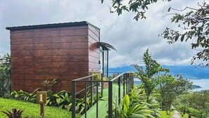 A wooden building with a glass door and a metal gate, surrounded by lush greenery and a pathway leading to a scenic view. -  Volcano and Lake View, Eco Glam #1. (La Fortuna)