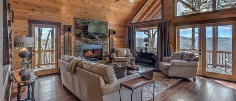 A cozy cabin interior with a fireplace, wooden floors, and large windows offering scenic views.