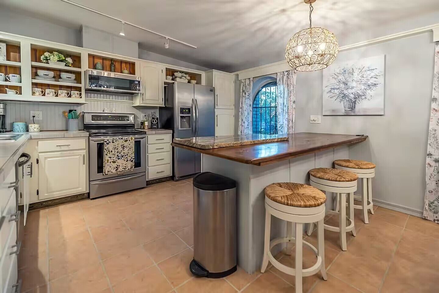 Une cuisine moderne avec des meubles blancs, des appareils en acier inoxydable, un îlot central doté de tabourets et un lustre décoratif.