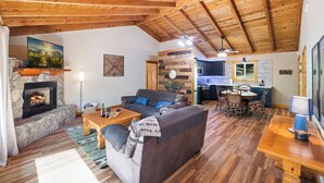 A cozy living room with a stone fireplace, a comfortable sofa, a wooden dining table, and a kitchen area in the background. - Blue Deer Mountain - Hot Tub | Fire Pit | Tree House Fun (Blue Ridge)