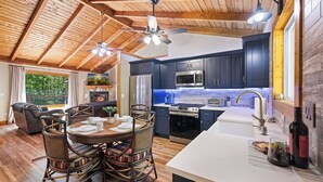 A modern kitchen with dark blue cabinets, stainless steel appliances, and a large island. There is a dining area with wicker chairs and a table set for four. - Blue Deer Mountain - Hot Tub | Fire Pit | Tree House Fun (Blue Ridge)