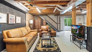 A modern living room with a leather sofa, armchair, and a coffee table. There is a staircase leading to another level, and a television on a stand. - Blue Deer Mountain - Hot Tub | Fire Pit | Tree House Fun (Blue Ridge)