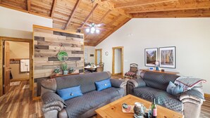 A cozy living room with two sofas, a coffee table with snacks and drinks, wooden ceiling beams, and framed pictures on the wall. - Blue Deer Mountain - Hot Tub | Fire Pit | Tree House Fun (Blue Ridge)