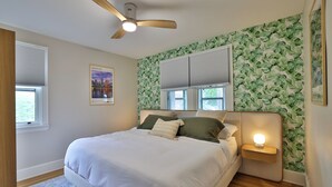 A bedroom with a bed, a ceiling fan, a nightstand with a lamp, and a window with blinds.