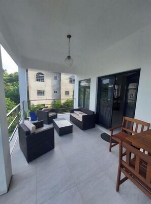 A balcony with outdoor seating, a dining table, and a view of a building with multiple windows. - Relax and Reconnect: A peaceful holiday home nestled in lush green surroundings. (Upton,  Ocho Rios)