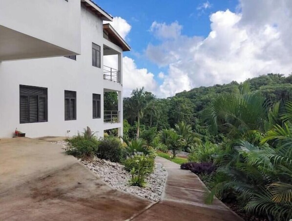 A two-story white building with a balcony, surrounded by lush greenery and a hilly landscape. - Relax and Reconnect: A peaceful holiday home nestled in lush green surroundings. (Upton,  Ocho Rios)