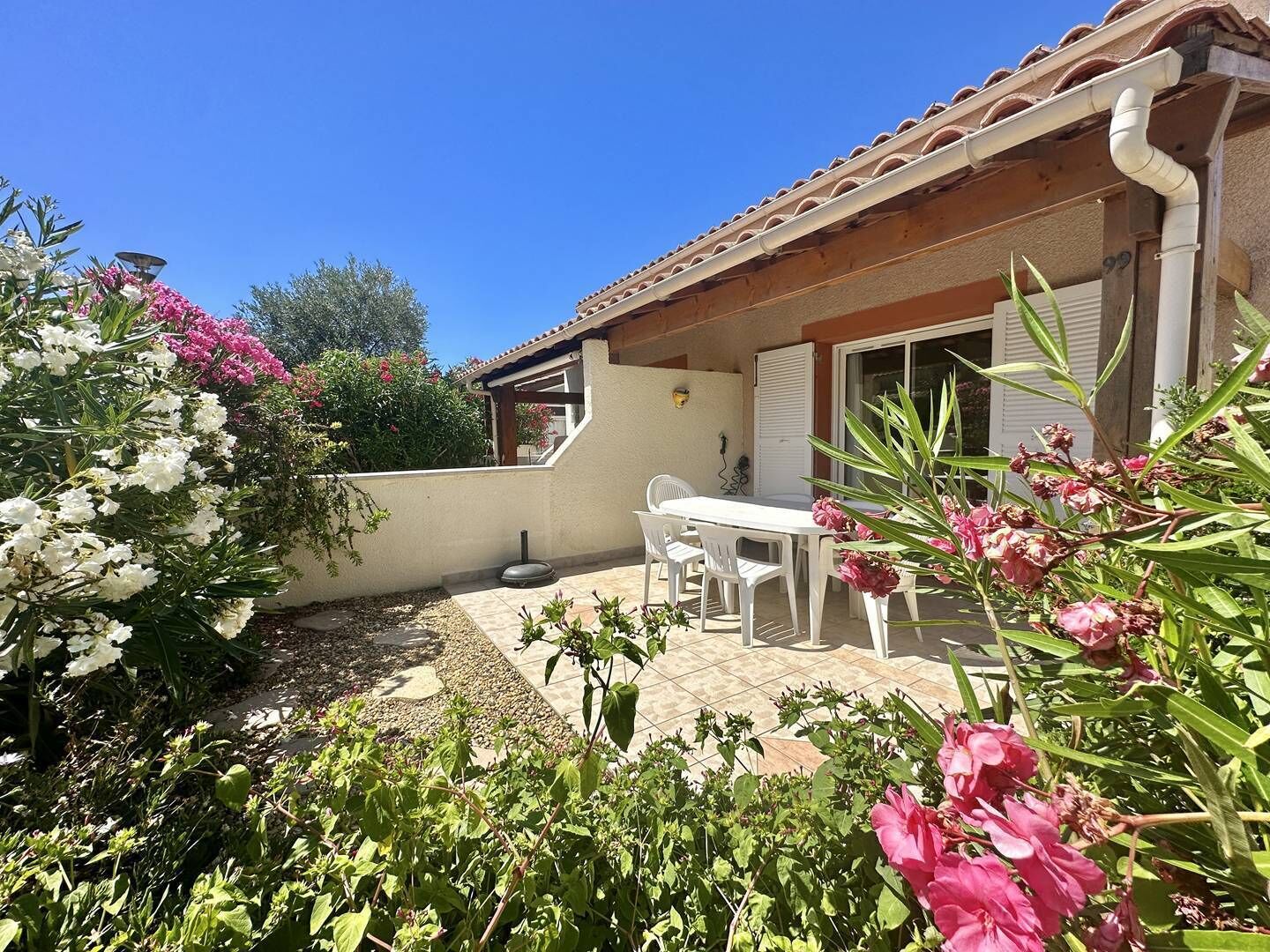 Une terrasse avec une table et des chaises blanches, entourée de plantes en fleurs et dotée d’un sol carrelé.
