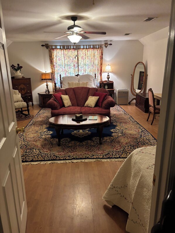 A living room with a red sofa, a coffee table, a ceiling fan, a bed with white linens, a dresser, a mirror, and a patterned rug.