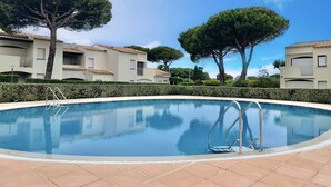 Une piscine à l'eau d'un bleu limpide, entourée d'une terrasse carrelée. On aperçoit des arbres et des bâtiments au fond.