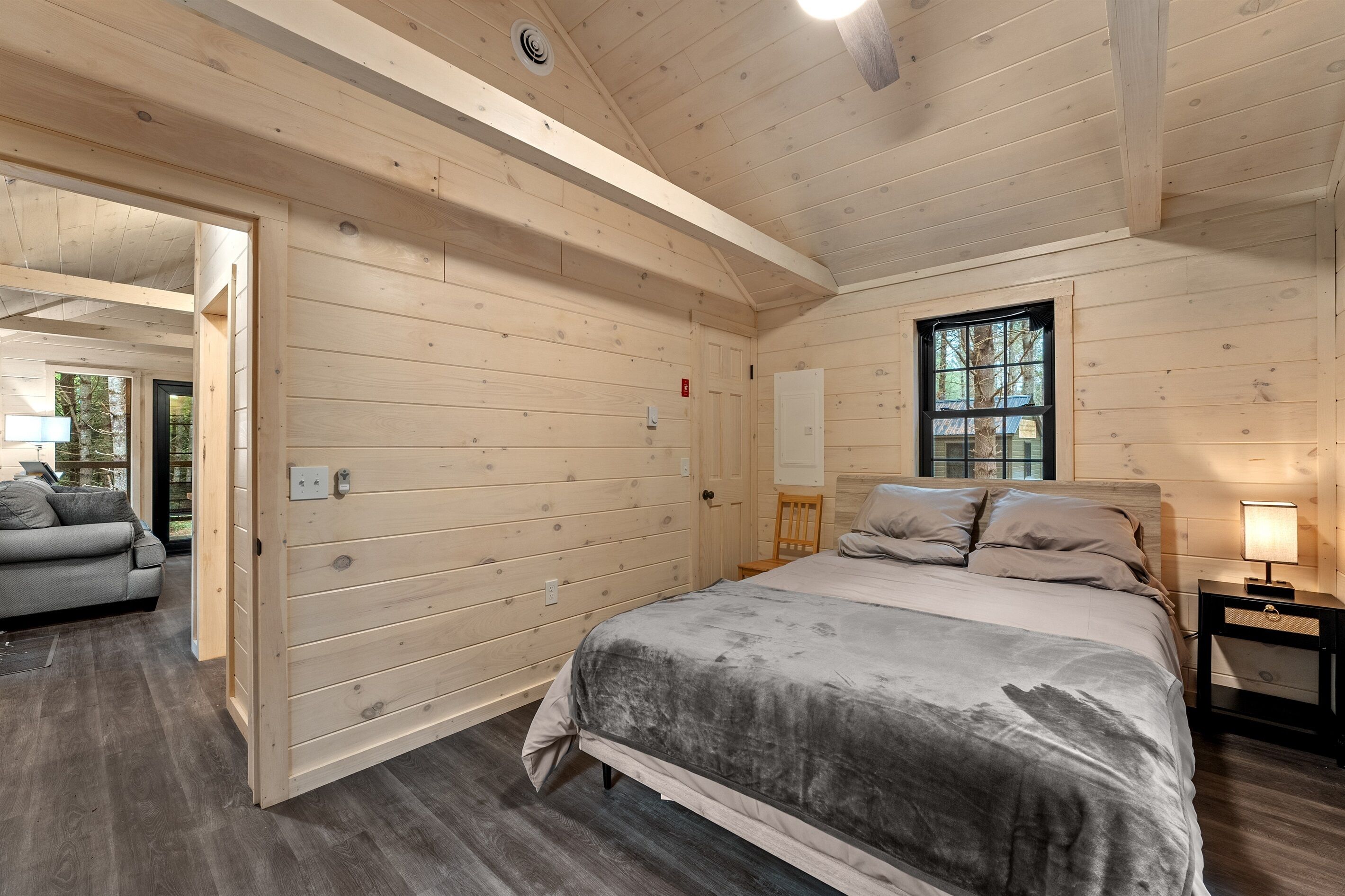 Un dormitorio de cabaña de madera con cama, mesita de noche, ventana y puerta que da a otra habitación.