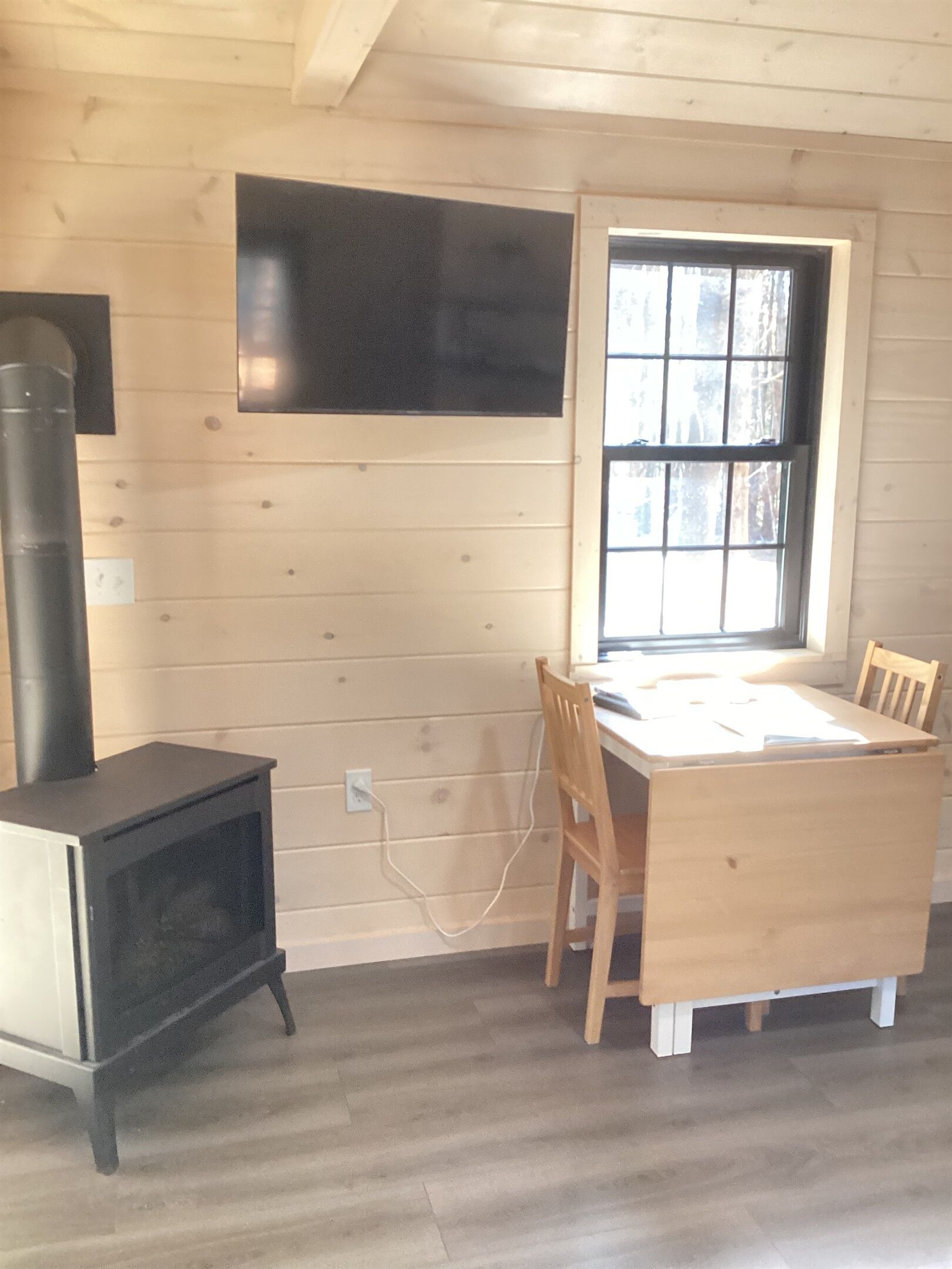 Een kamer met houten inrichting, een zwarte kachel, een tafel met stoelen en een raam met uitzicht op bomen.
