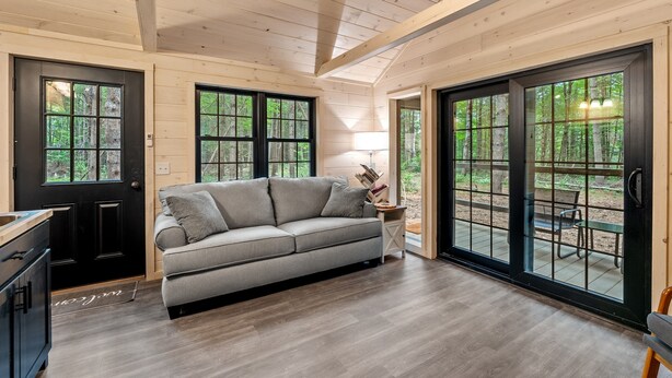A modern log cabin interior with a gray sofa, wooden flooring, and large windows offering a view of the surrounding forest.