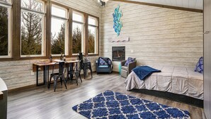 A cozy room with a wooden ceiling, large windows, a fireplace, a bed, and a dining area with chairs and a table.