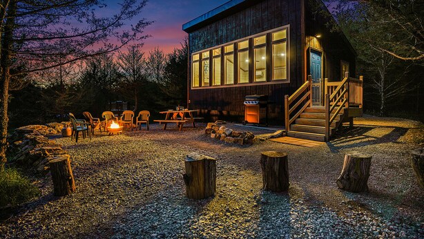 A rustic cabin with a large windowed front, a wooden deck, and a fire pit area.