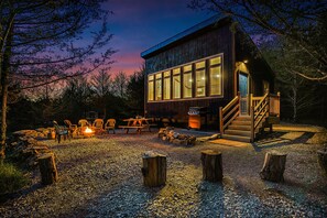 A rustic cabin with a large windowed front, a wooden deck, and a fire pit area. - Modern Cabin. Communal Sleeping - 2 Queen Beds + Full Trundle (Ashland)