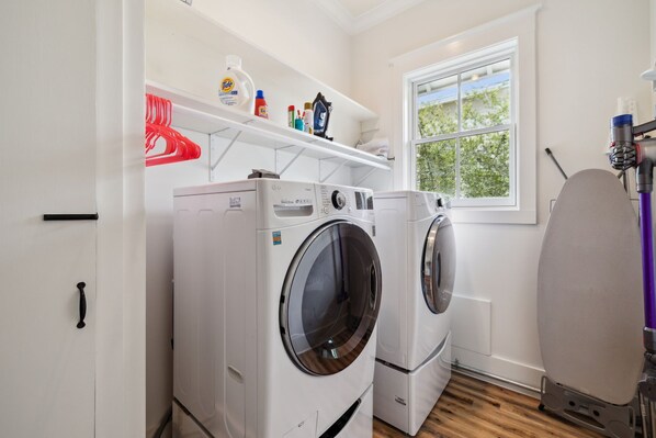 A laundry room with two front-loading washing machines, a shelf with laundry supplies, and a window with a view of trees. - Pet Friendly, Lake District Watercolor, Tesla Charger (Santa Rosa Beach)