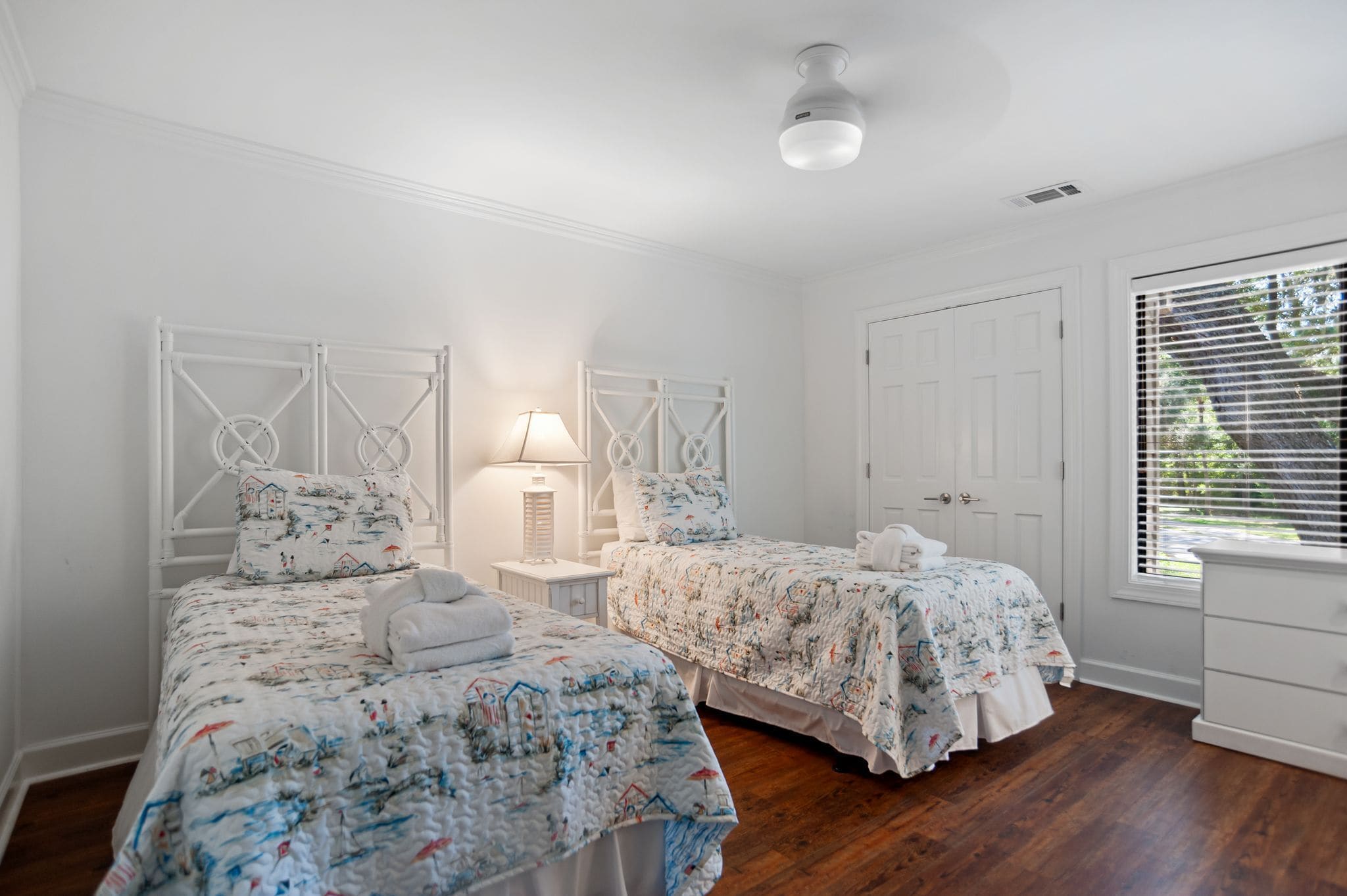 A bedroom with two beds, white walls, wooden flooring, a ceiling light, and a window with blinds.