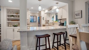 A modern kitchen with white cabinetry, a marble island with bar stools, pendant lighting, and a view into the dining area. - 2426 Inland Harbour at The Sea Pines Resort (Hilton Head Island)