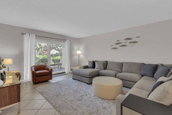 A modern living room with a grey sectional sofa, a round ottoman, a brown leather chair, and a wooden side table. - 2264 Heritage Villas at The Sea Pines Resort (Hilton Head Island)