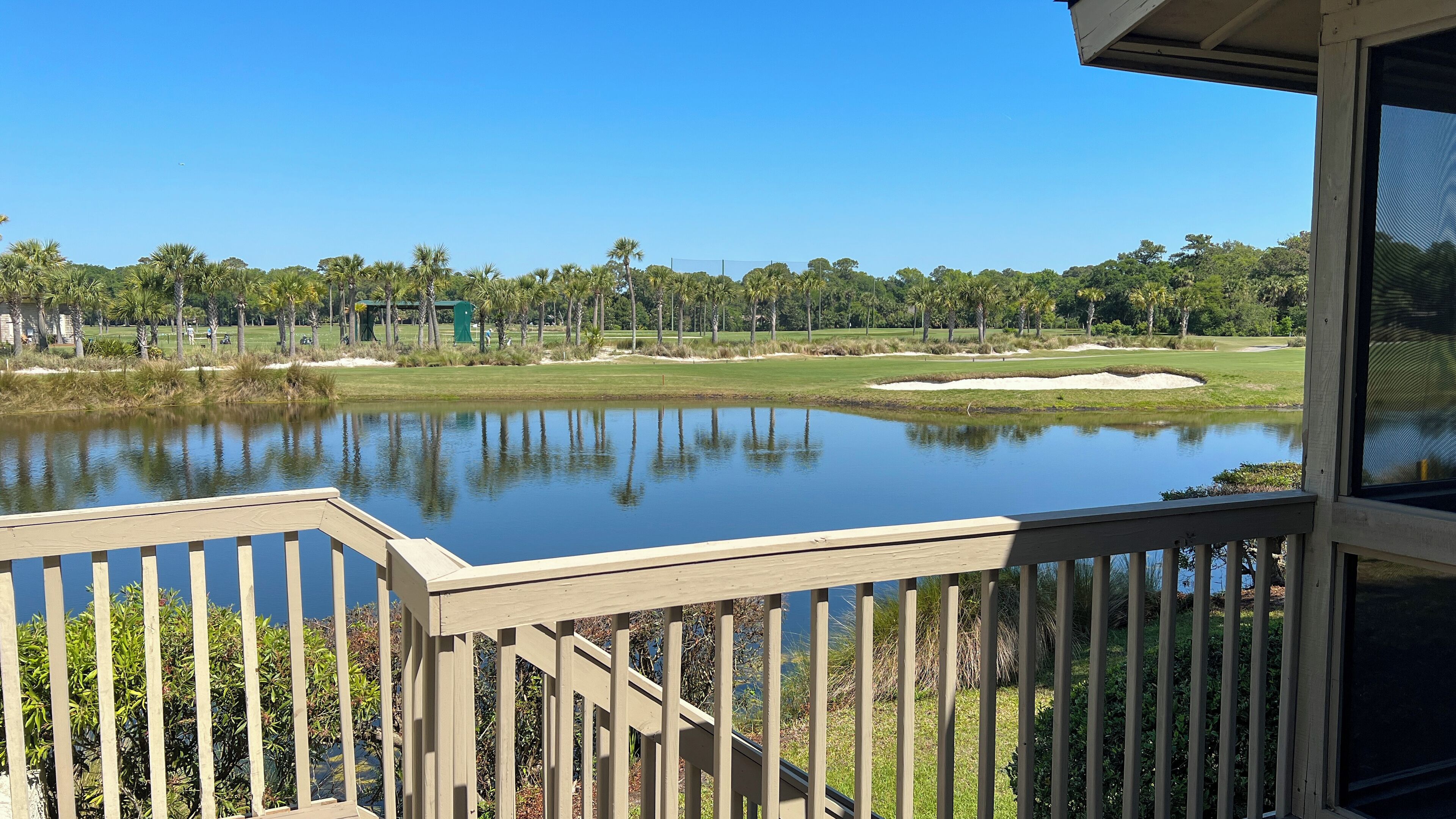 A view from a deck overlooking a golf course with a pond, palm trees, and a clear blue sky.