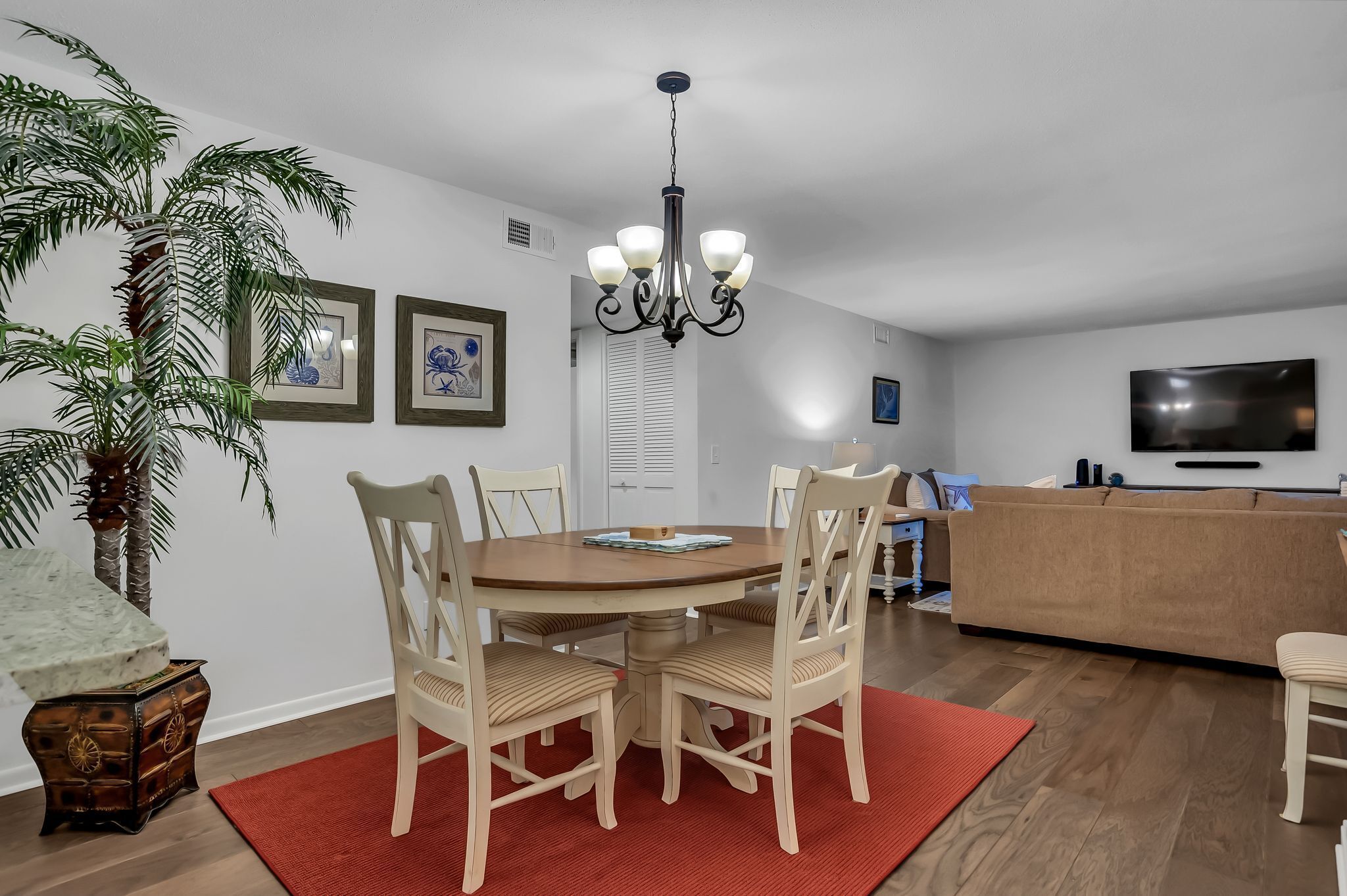 A dining area with a round table, four chairs, a chandelier, a large potted plant, and a red rug.