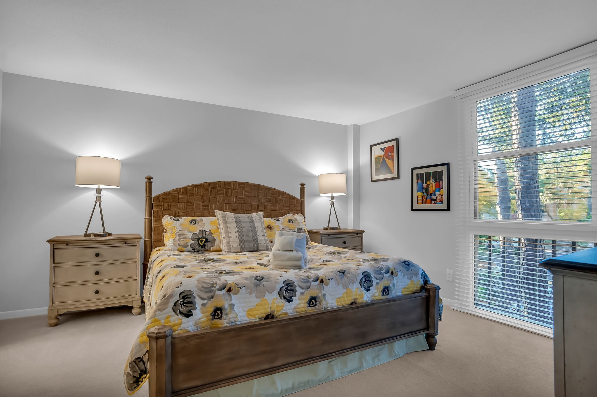 A bedroom with a wooden bed, two bedside tables, a large window with blinds, and a wall-mounted picture.